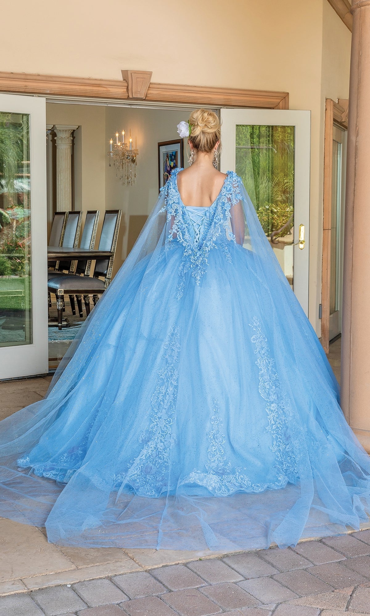 A woman in the Quinceanera Dress 1678 by Dancing Queen, a light blue floor-length A-line gown with lace details and removable draped sleeves, stands with her back to the camera facing a dining room doorway.